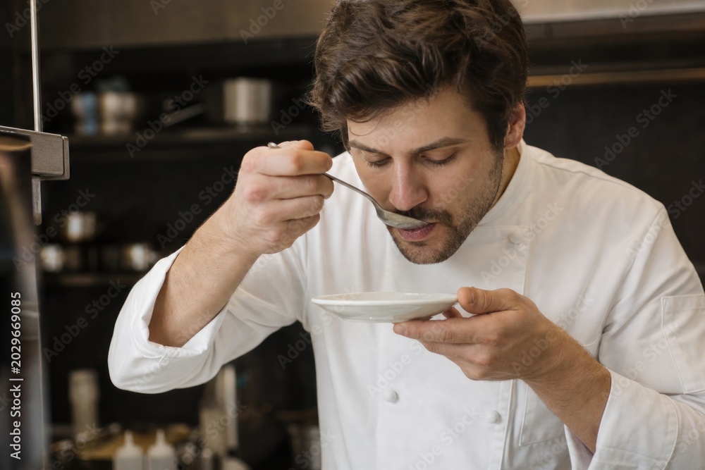 Fototapeta Professional chef in white uniform meticulously tasting fine sauce from small spoon in modern kitchen, culinary expertise, food preparation, gastronomy, close-up detail, warm lighting, restaurant