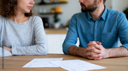Unrecognizable couple silently sitting at kitchen table with utility bills spread between them. Concept of financial crisis in relationships and emotional pressure caused by debt.