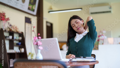 young Asian woman stretching neck shoulder relaxing from remote work using laptop computer at home cafe office desk wellness break during online business