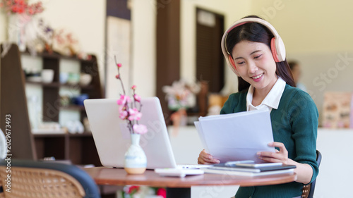 young Asian women wearing headphones working from home cafe using laptop computer reading document paper remote work online business freelance study learning