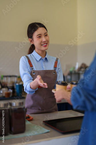 young Asian female barista cafe owner serving customer greeting welcome gesture coffee shop small business hospitality service industry counter friendly smile