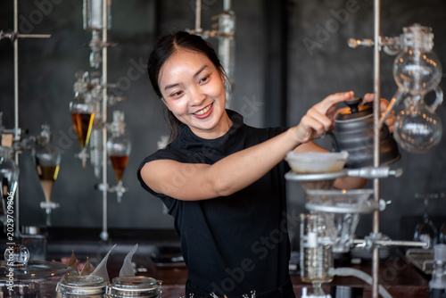 Bild auf Leinwand Smiling Asian woman pouring hot water from a kettle onto a coffee drip filter integrated into a glass apparatus setup