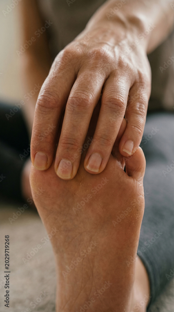 Fototapeta premium Extreme close-up of woman hand holding bare foot while stretching at home, female preparing for fitness training and muscle warm-up in minimalist living room