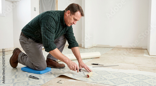Man installing patterned linoleum sheet flooring. Male handyman kneeling on a foam pad and using a seam roller. Home interior renovation and remodeling project