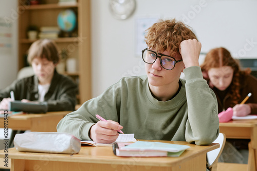 Portrait of Caucasian teenager boy studying at school desk, writing notes and showing learning stress during class