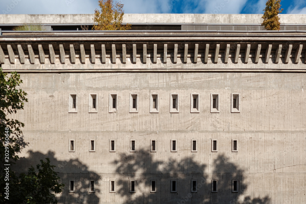 Obraz premium A flat, front-on architectural shot of a massive brutalist concrete wall featuring a rhythmic, repeating pattern of small vertical windows and structural facade details under a blue sky.