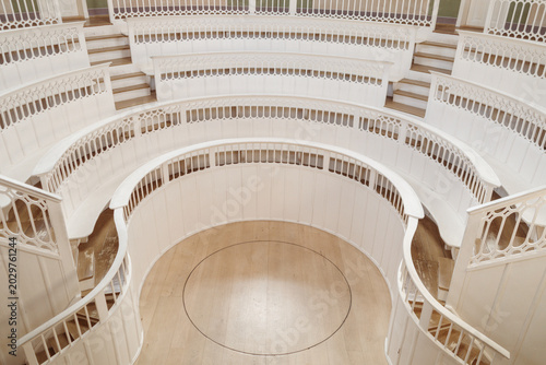 An indoor, high-angle photograph looking down into an empty, historic anatomical theater, revealing concentric rings of steep, off-white wooden seating surrounding a central wooden floor with a circul