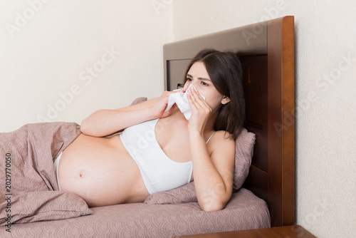 Картината върху платно Portrait of young unhappy pregnant woman leaning on pillows on bed and blowing her nose into tissue