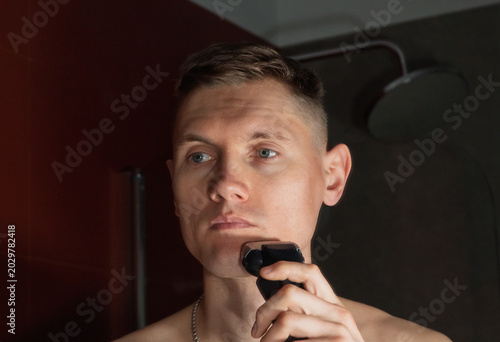 Foto Attractive European-looking man shaves with an electric razor in front of a bathroom mirror
