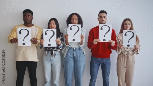 Five young adults from different backgrounds stand in a modern studio holding large question mark signs. They look towards the camera, showcasing curiosity and engagement in their activity.
