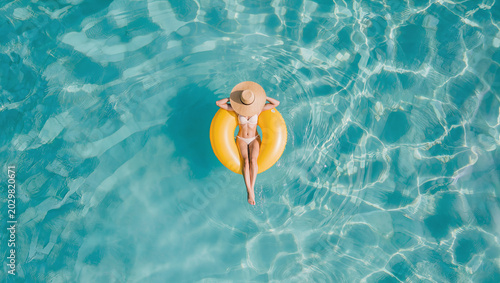 Overhead view of a person relaxing on a yellow inflatable ring in clear turquoise water, wearing a bikini and straw hat in a calm summer setting.