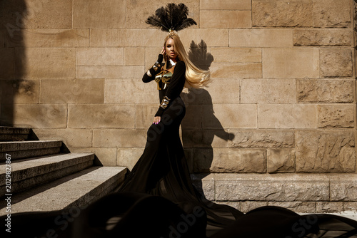Billede på lærred Majestic female model in avant-garde flowing black gown with metallic gold armor and elaborate feather headdress posing against a textured stone wall in a high-fashion fantasy editorial