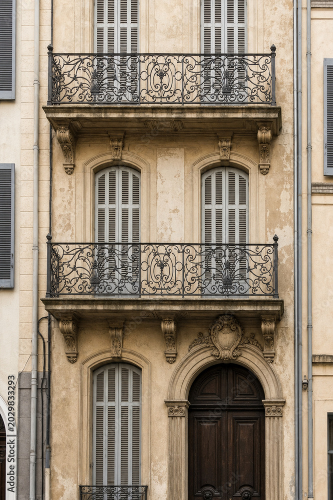 Obraz premium Close-up of a historic building facade with ornate wrought-iron balconies, arched entry, and sunlit beige stone, featuring symmetrical windows and classical architectural details.