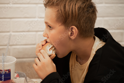 Kid teenager boy sits at a fast food restaurant, eating burger and drinking soda
