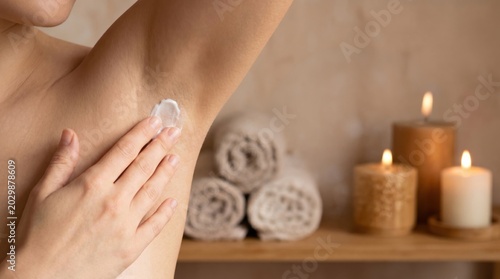 A person applies cream to their underarm, with rolled towels and lit candles in the background creating a calming, spa-like atmosphere.