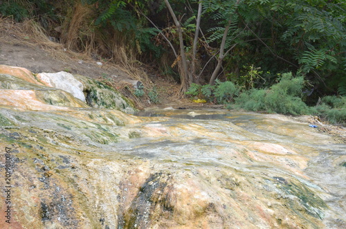 A detailed view of colorful mineral terraces formed by thermal spring waters. The rocky surface shows yellow, orange, and green textures from salt deposits near wild hot baths in the Caucasus