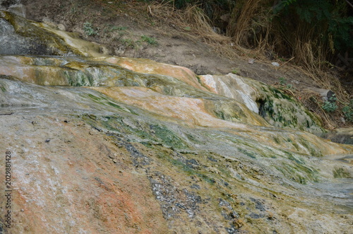 A detailed view of colorful mineral terraces formed by thermal spring waters. The rocky surface shows yellow, orange, and green textures from salt deposits near wild hot baths in the Caucasus