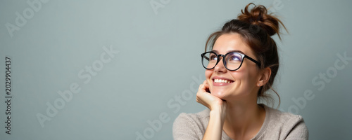 Young woman with glasses smiles, looking up thoughtfully. She rests chin on hand, thinking about new ideas. Casual indoor studio portrait with copy space.