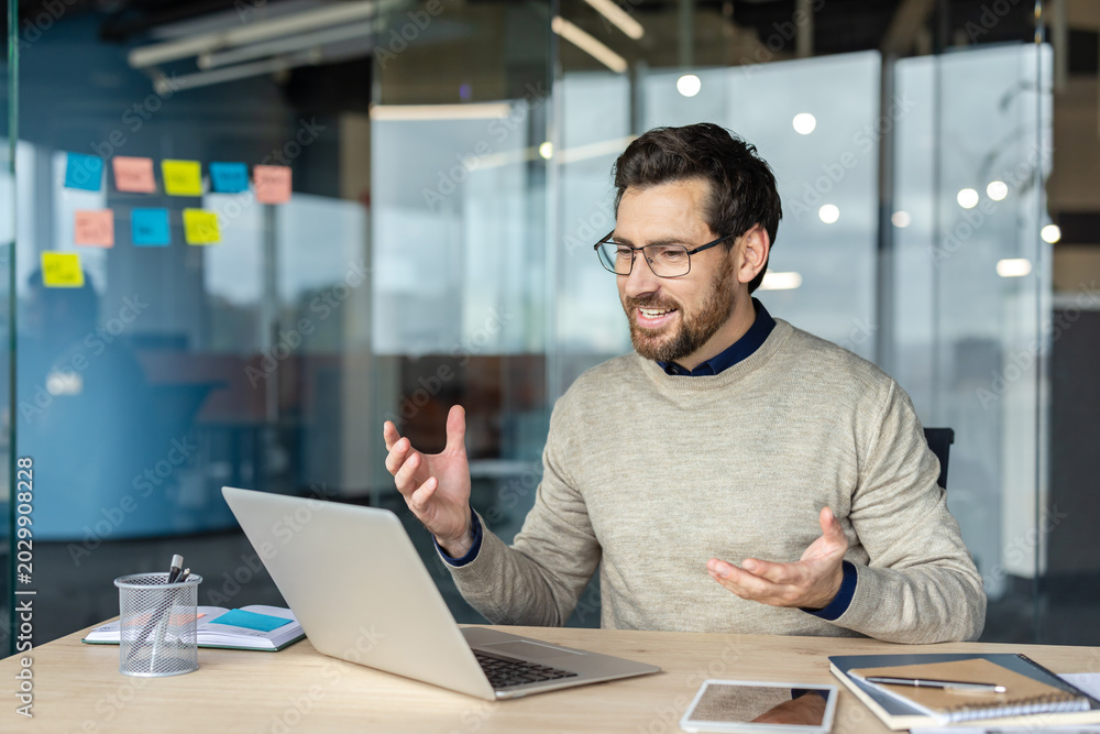 Obraz premium Bearded man wearing glasses engaging in a video call and gesturing while communicating on a laptop from his modern office workspace, showcasing remote work and online connection
