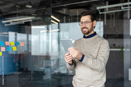 Cuadro en lienzo Smiling man standing in a modern office, wearing glasses and a sweater, holding