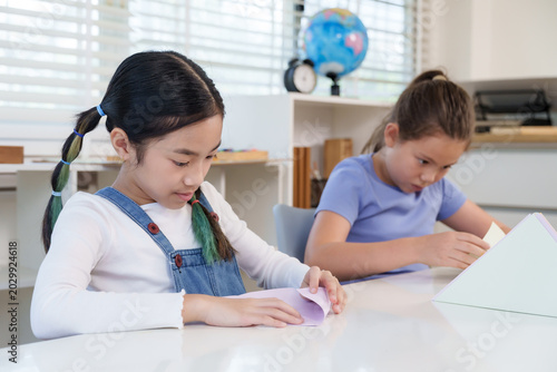 Bild auf Leinwand Asian preteen girl folds colored paper carefully while caucasian classmate conce