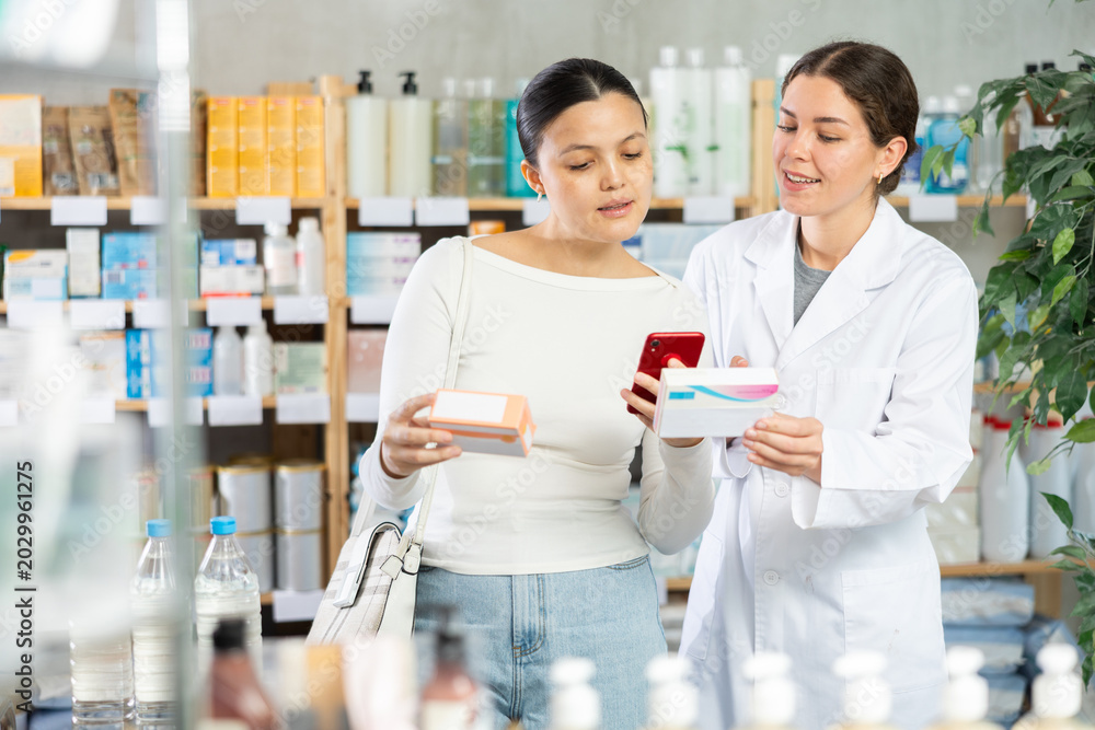 Fototapeta premium Attentive young girl pharmacist advising asian female client choosing and scanning qr code for box of pills in modern pharmacy store