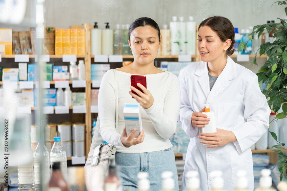 Fototapeta premium Interested Asian female customer scanning QR code on package of body cream with phone to confirm product safety, while smiling young pharmacist suggesting additional options in pharmacy