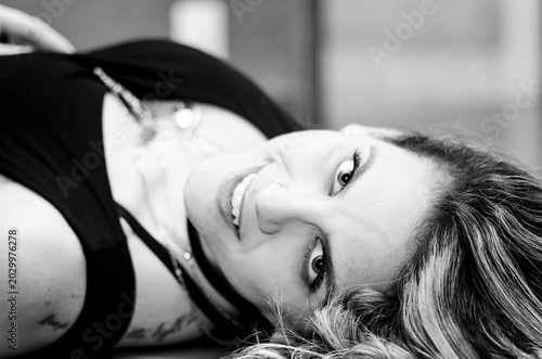 Black and white portrait of a woman lying down, smiling directly at the camera. The close-up framing emphasizes her gaze. The image conveys intimacy, lightness, and naturalness.