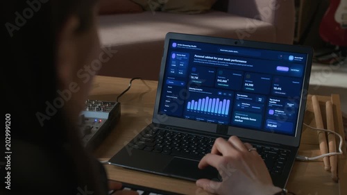 Over shoulder shot of female producer monitoring streaming analytics on music selling platform while using touchpad on laptop in home recording studio