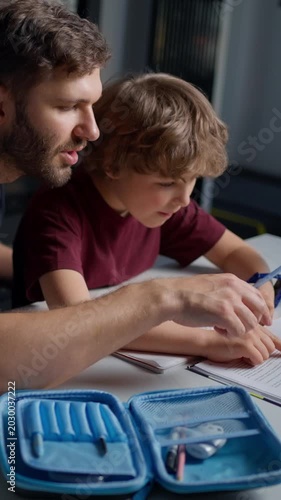 A dedicated father and his young son engaged in home study session, focusing on writing and reading together at a table with educational supplies and stationery visible