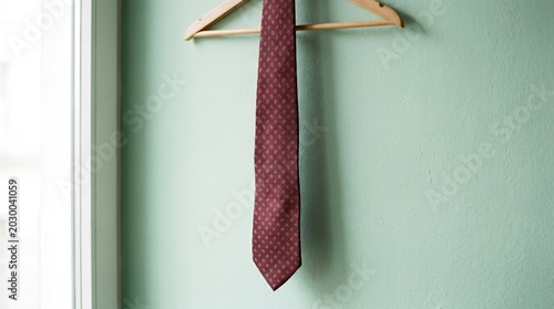 Burgundy patterned necktie hanging on wooden hanger against green wall.