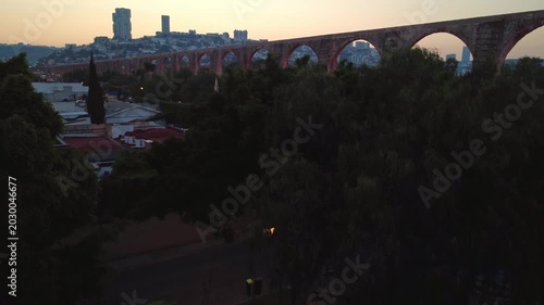 Cinematic tilt up and pan aerial movement revealing Aqueduct of Queretaro above dense trees and urban skyline in Santiago de Queretaro Mexico during sunset