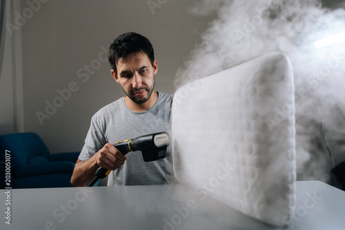 Young adult man steam-cleaning white pillow using handheld steamer, removing allergens, dust mites and germs for deep-cleaned, fresh bedding and improved home hygiene.