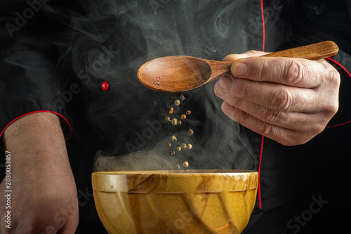 A chef stands in a kitchen, using a wooden spoon to add spices to a bowl. Steam rises around while the chef prepares a dish. The scene shows cooking activity in a vibrant atmosphere