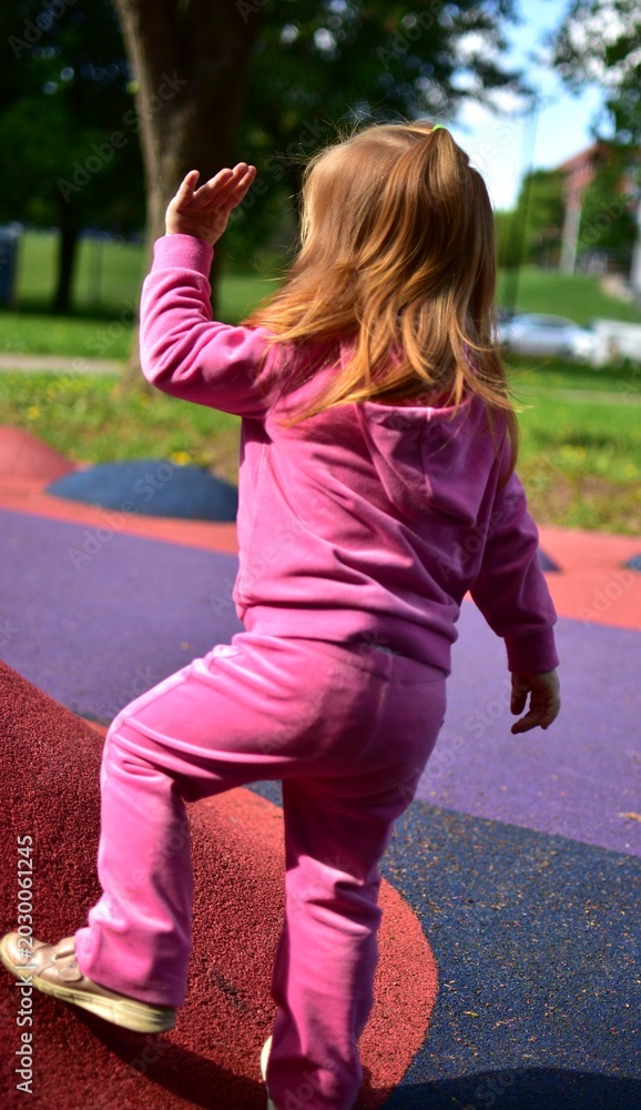 Fototapeta premium Toddler girl walking and balancing on colorful playground surface in sunny outdoor park. Active childhood, coordination training and playful exploration concept with modern recreational equipment.
