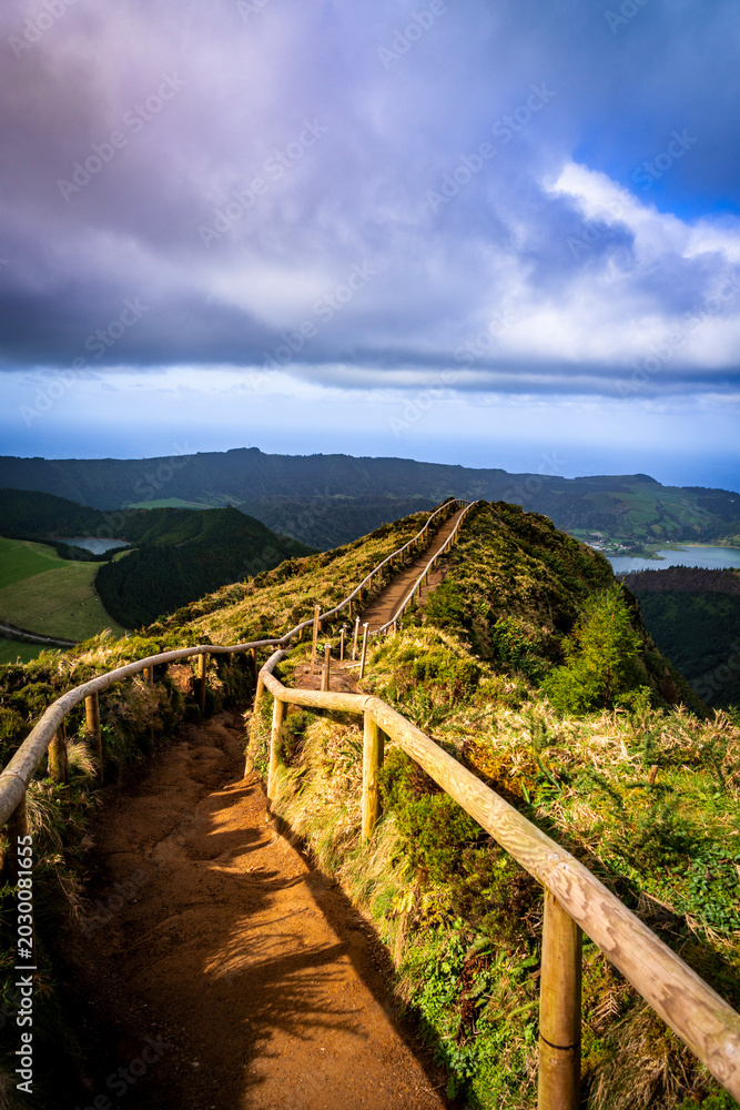 Fototapeta premium Wooden-railed hiking trail winding along a lush volcanic ridge in the Azores with crater lakes and ocean views under a dramatic cloudy sky.
