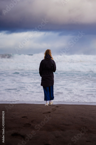 Blonde woman in a black jacket standing alone on a dark sand beach facing powerful crashing waves under a dramatic stormy cloudy sky.