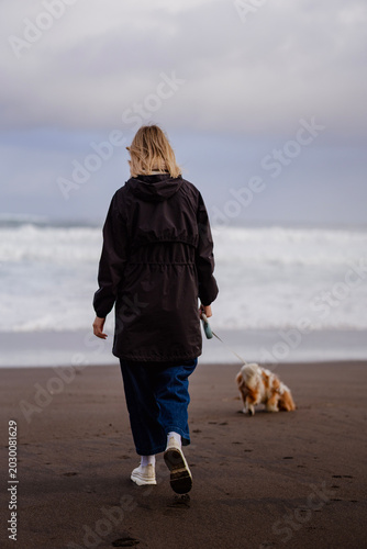 Blonde woman in a black rain jacket walking her Cavalier King Charles Spaniel on a dark volcanic sand beach with stormy waves in the background.