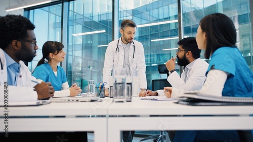 Head doctor standing and addressing diverse medical team in bright meeting room, colleagues actively listening and engaging. Morning briefing starting with leadership guidance.