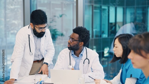 Indian and African American male doctors having discussion around laptop, Asian woman listening, others working. Medical team sharing opinions during collaborative meeting. Multiethnic medics.
