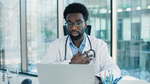 African American male doctor explaining while working on laptop via video call on web camera, wearing glasses, stethoscope. Medical professional speaking during remote consultation or virtual meeting.