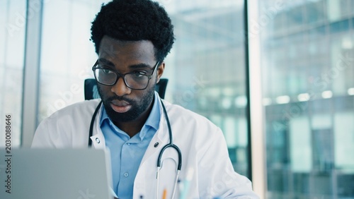 Handsome young African American male doctor focused on laptop, wearing glasses and stethoscope, working in bright office. Medical professional reviewing patient data or digital records.