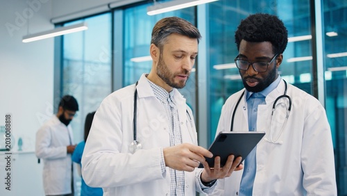 Foto Caucasian male doctor pointing at digital tablet held by African American colleague during discussion in office, both wearing lab coat and stethoscope, collaborating closely