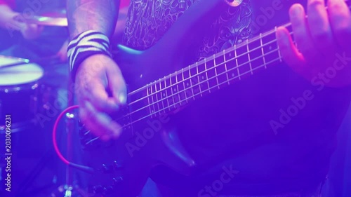 Close up of musician's hands playing bass guitar during punk rock show. Stage lights create colorful, hazy atmosphere. Drums in background. Energetic live music performance concept
