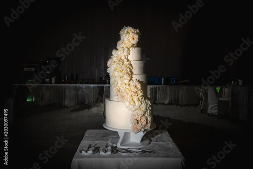 Elegant white wedding cake with cascading roses on a table at night, luxury wedding reception overlooking tropical beach background with champagne glasses.