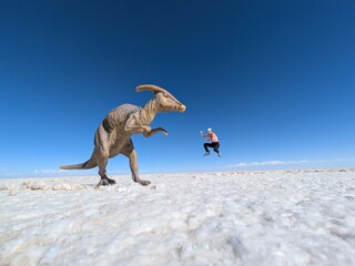 Playful forced perspective trick photography on a vast white salt flat featuring a toy dinosaur in the foreground and a happy female traveler jumping in the background under a clear blue sky.