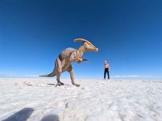 Playful forced perspective trick photography on a vast white salt flat featuring a toy dinosaur in the foreground and a happy female traveler jumping in the background under a clear blue sky.