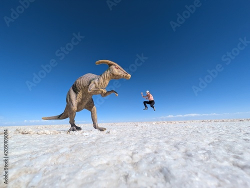 Obraz na plátně Playful forced perspective trick photography on a vast white salt flat featuring a toy dinosaur in the foreground and a happy female traveler jumping in the background under a clear blue sky