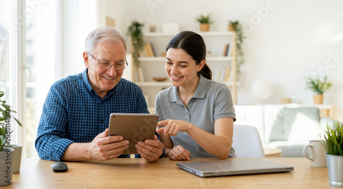 Young woman helping an elderly man with a tablet. Happy senior caucasian grandfather and granddaughter using technology together at home