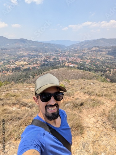 Cheerful man taking a selfie on top of a mountain overlooking a scenic valley town, enjoying a sunny day of hiking, adventure travel, outdoor recreation, freedom and active lifestyle.